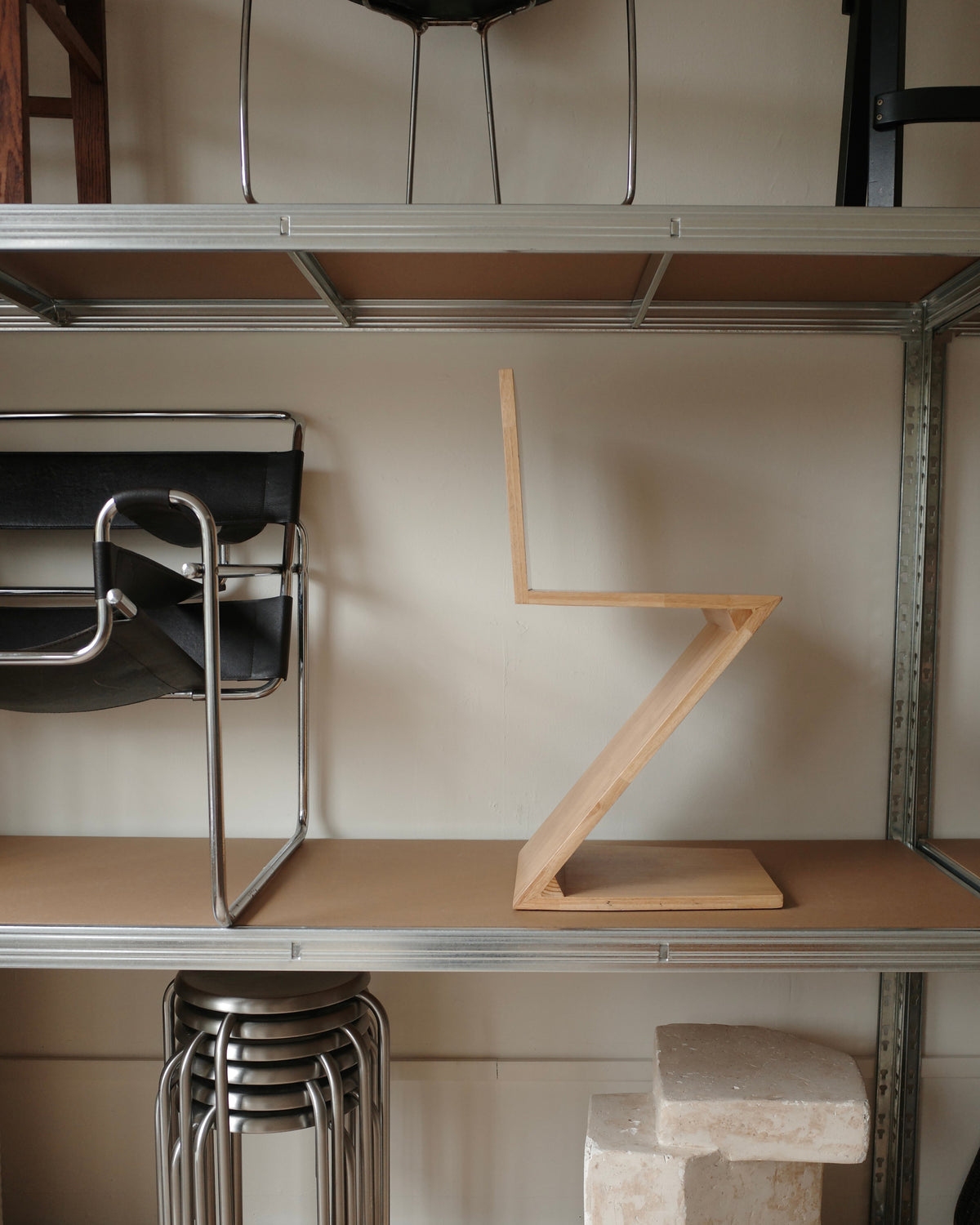A minimalist wooden Z-shaped shelf riser on a brown shelf of a metal modular storage rack, alongside other modern furniture pieces.