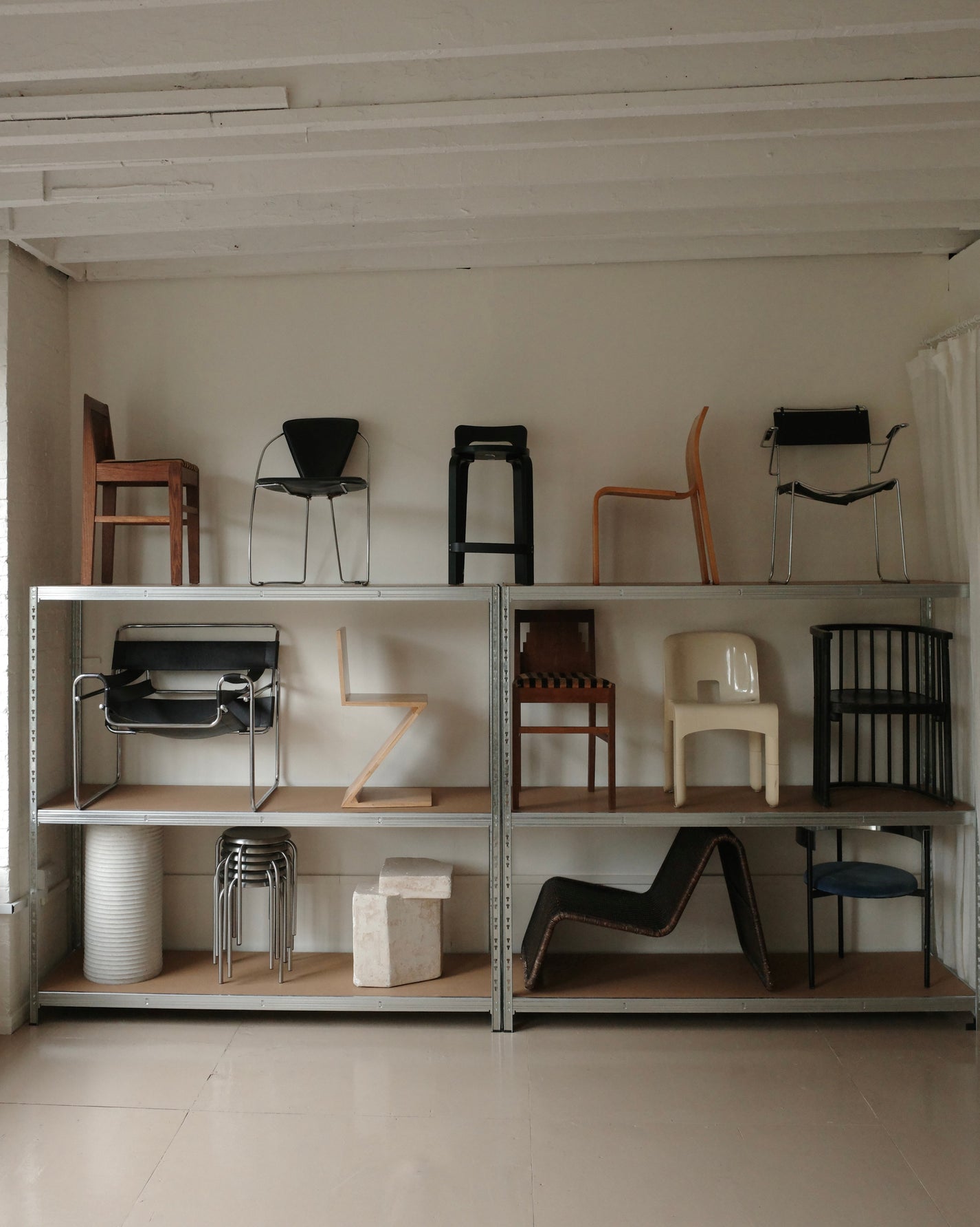 A collection of various chairs and stools displayed on a two-tier metal shelving unit in a minimalist room with white walls and ceiling beams.