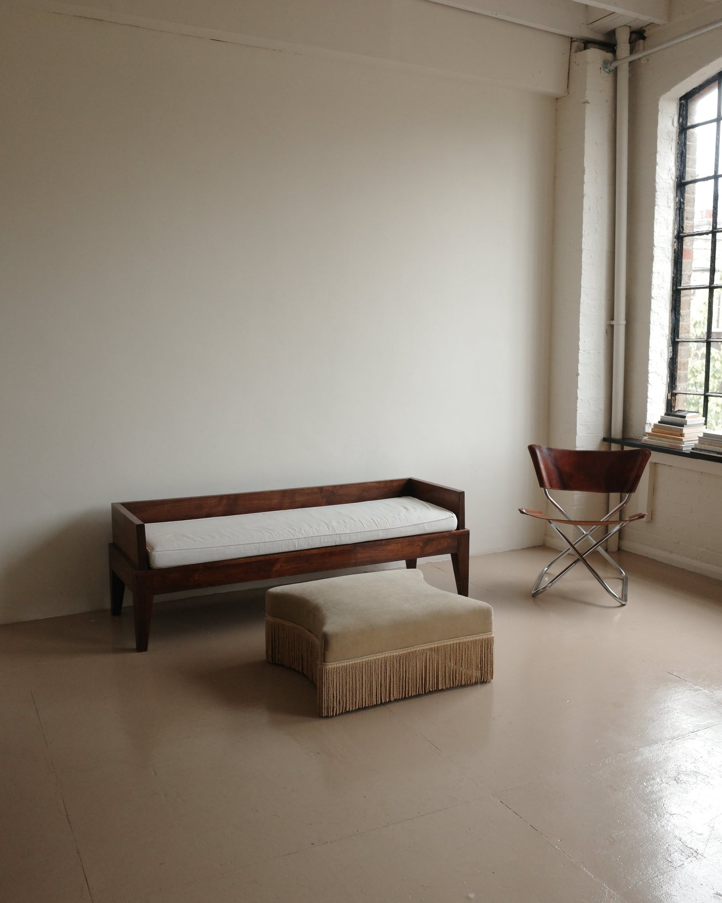 A minimalist room scene featuring a dark wood bench with a white cushion, a matching beige fringed ottoman, and a red folding chair near a window.
