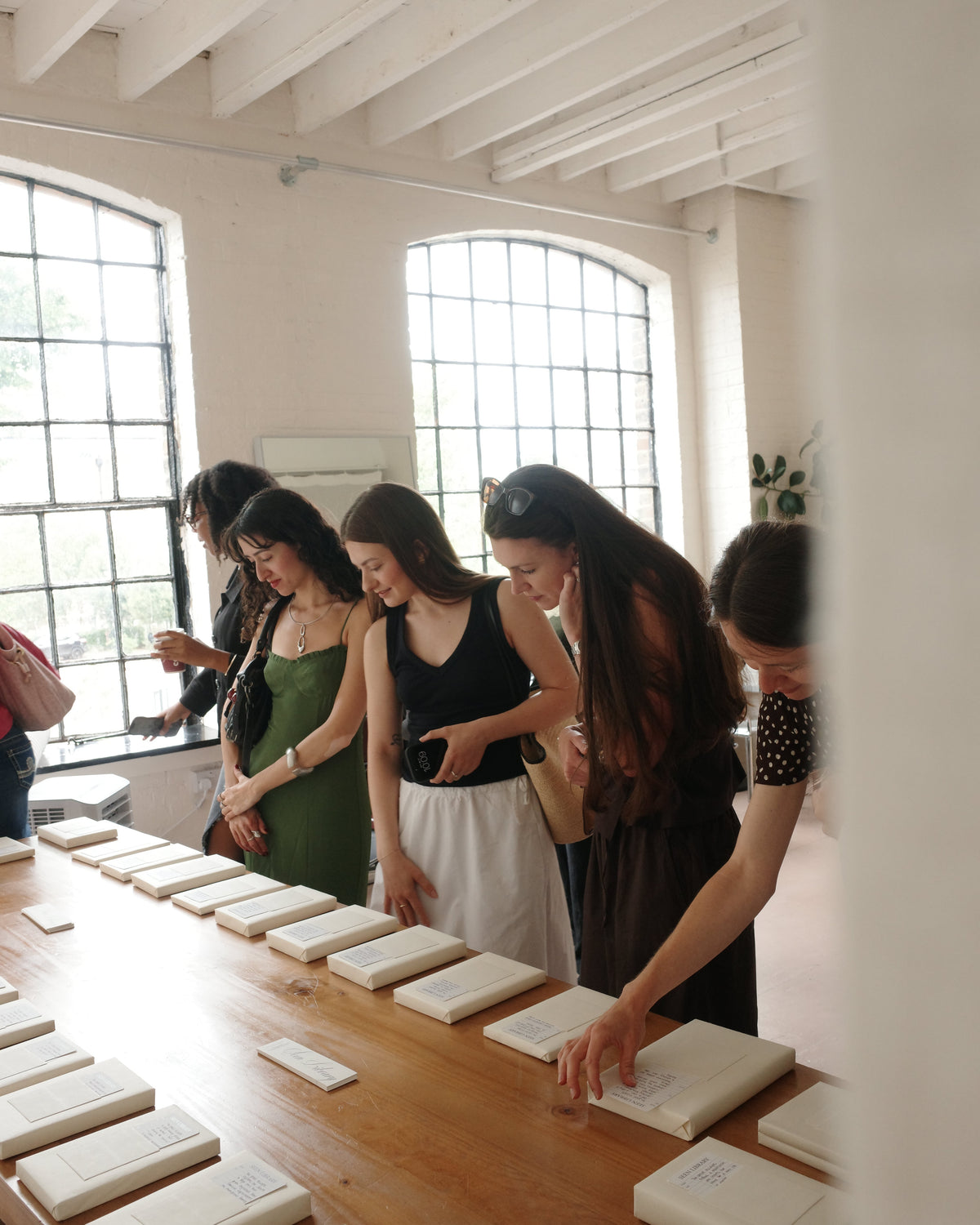 A group of women gathered around a long wooden table, examining small white booklets or cards laid out on the surface in a bright, white-walled room with large arched windows.