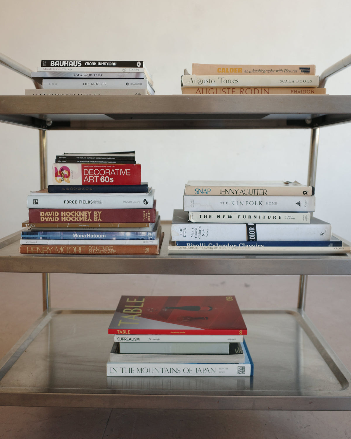 A mid-century modern metal bookshelf with three tiers, holding a curated collection of books. The bookshelf features a brushed silver/chrome frame with a glass bottom shelf.