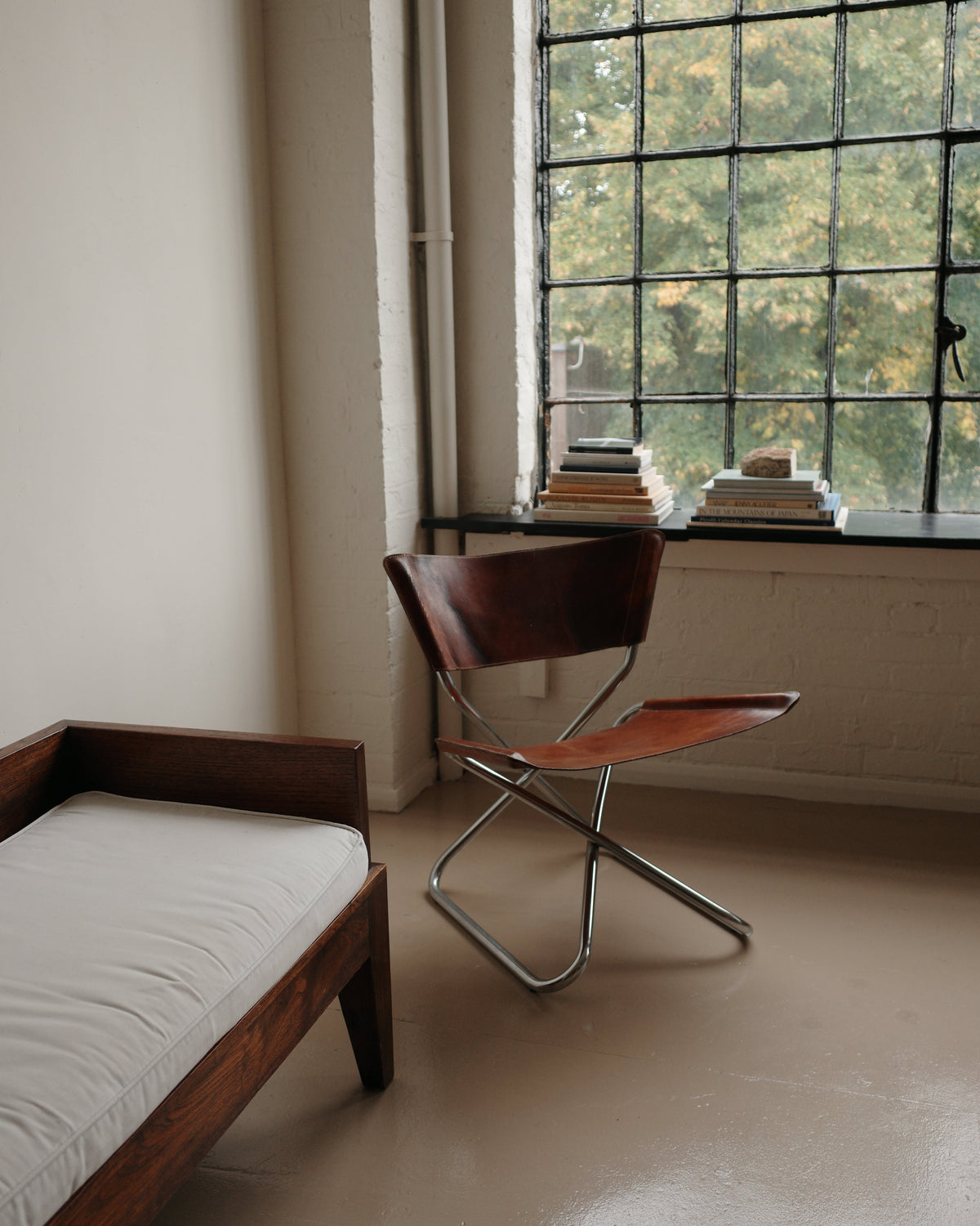 A mid-century modern folding chair with a dark brown wooden seat and backrest and a chrome metal frame, positioned next to a large multi-pane window in a minimalist room.